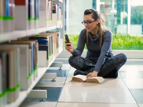 college student sitting on floor in library, typing on mobile